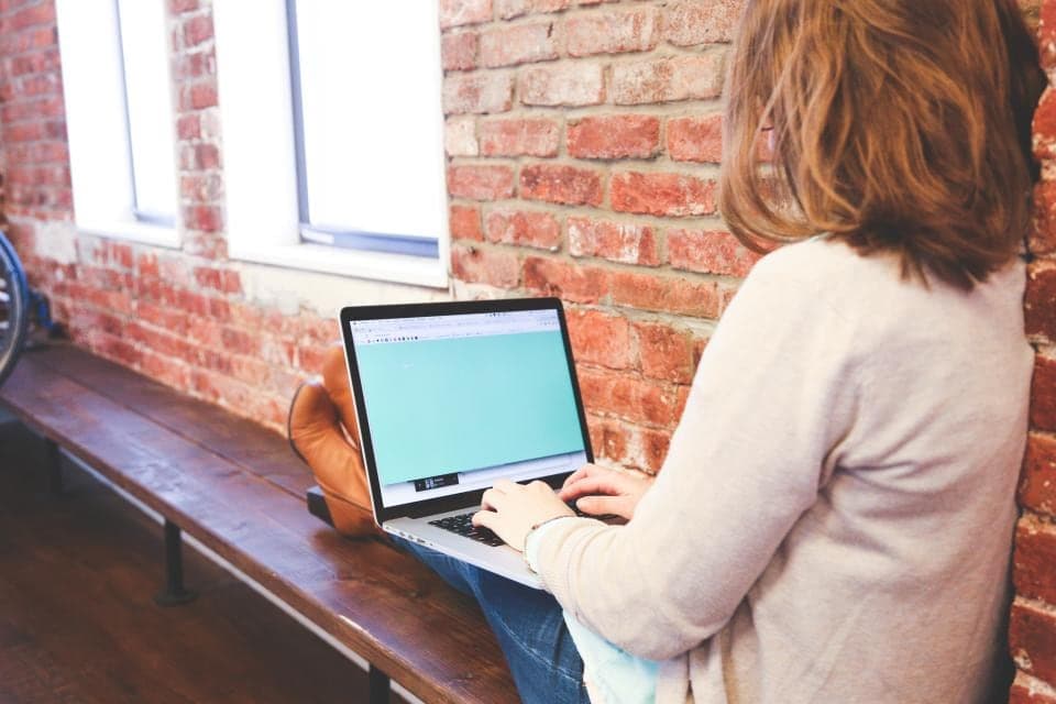 A developer working on a laptop at a wooden bench beside a brick wall and window