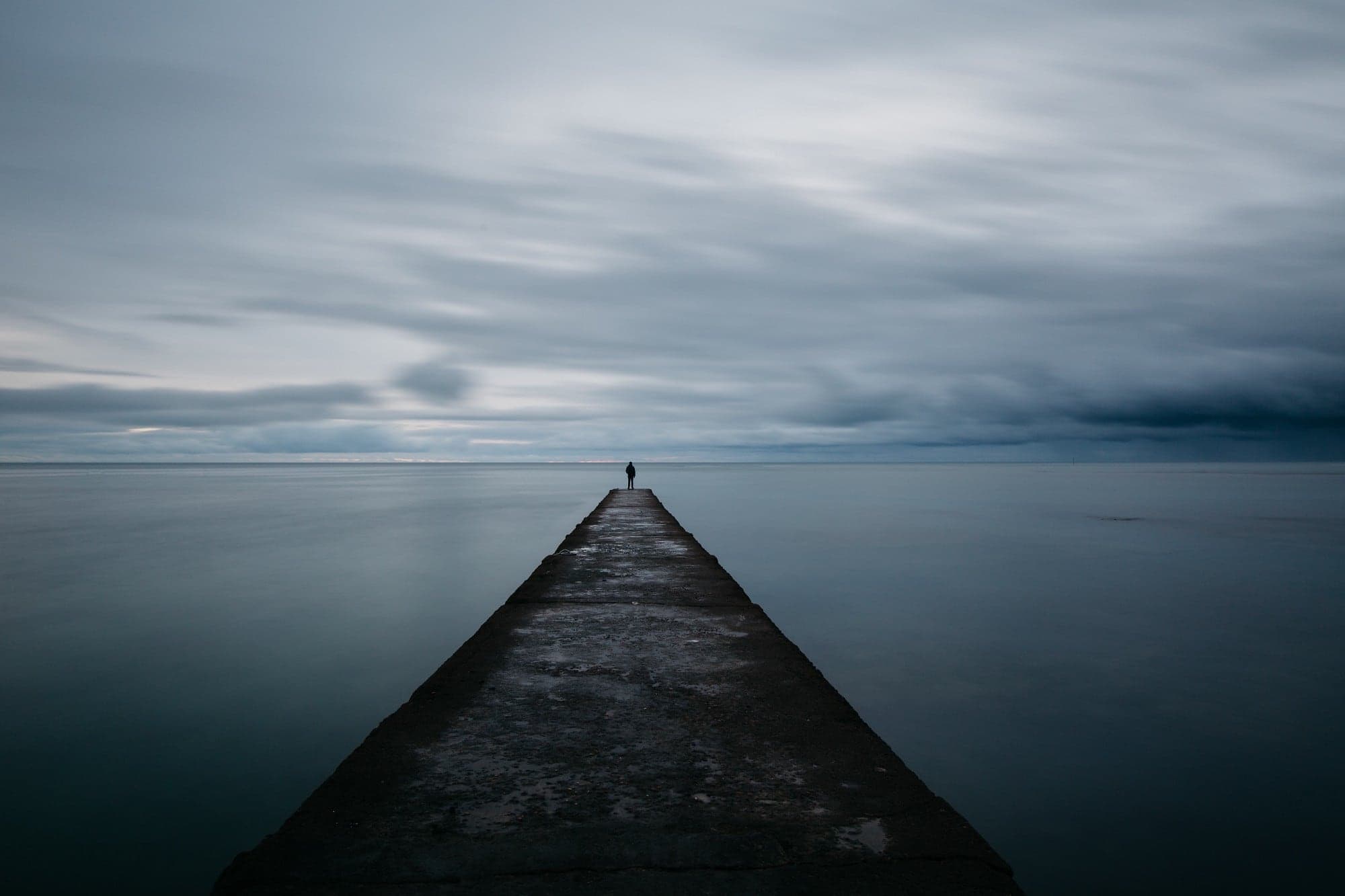 A lone figure at the end of a long stone jetty stretching into a calm grey sea