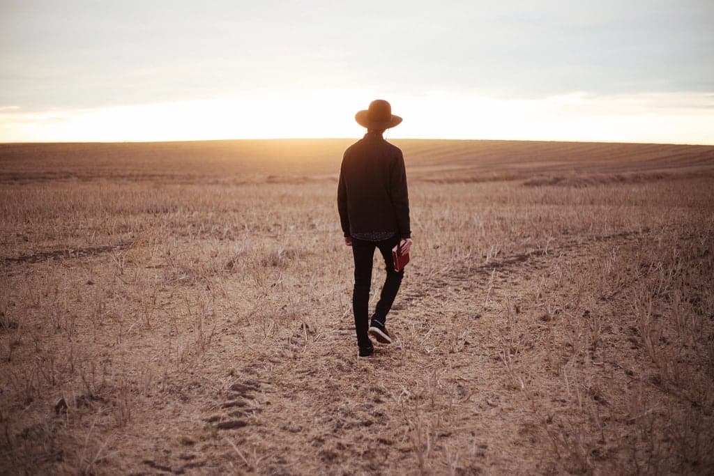 A person in a hat walking across an open field towards the low evening sun